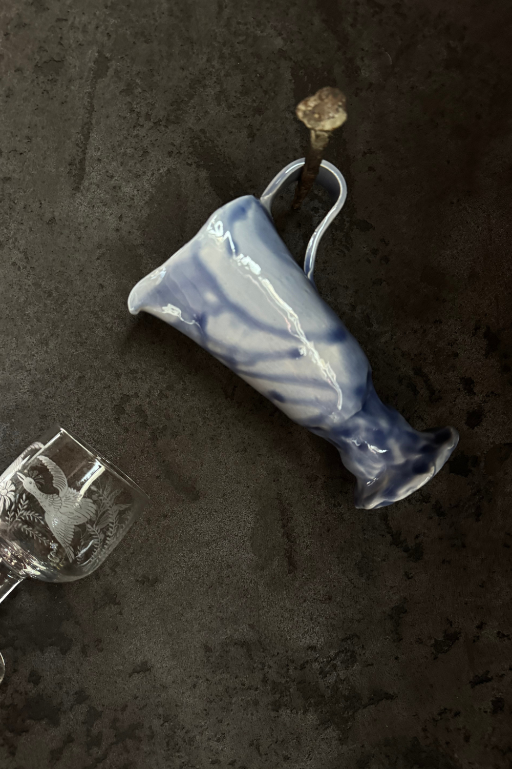A blue ceramic carafe resting on a textured surface, with an etched glass in the background.