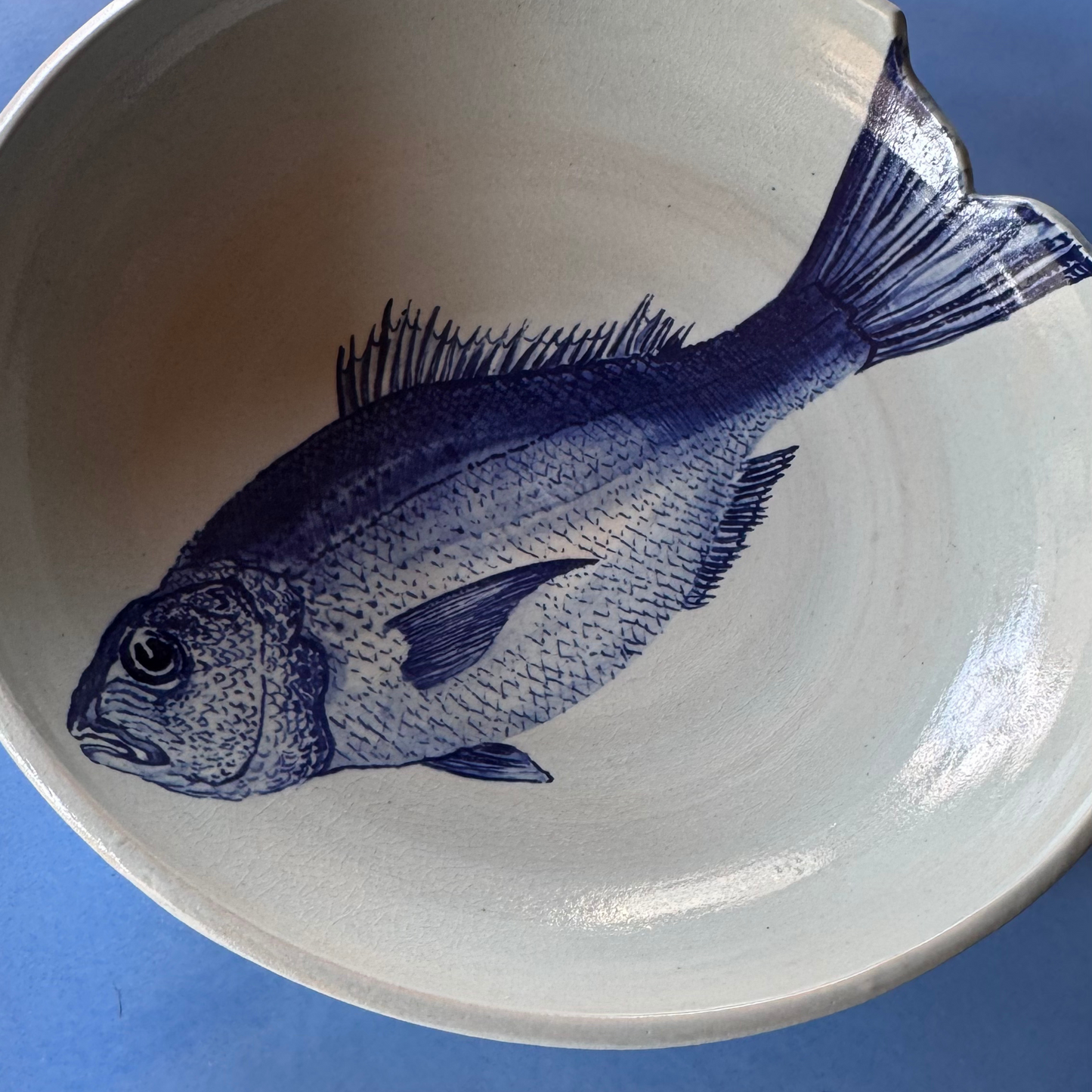 Handcrafted ceramic bowl featuring a detailed blue dorade fish illustration on a light, off-white glaze. The organic shape and hand-painted design give it a unique artisanal touch.