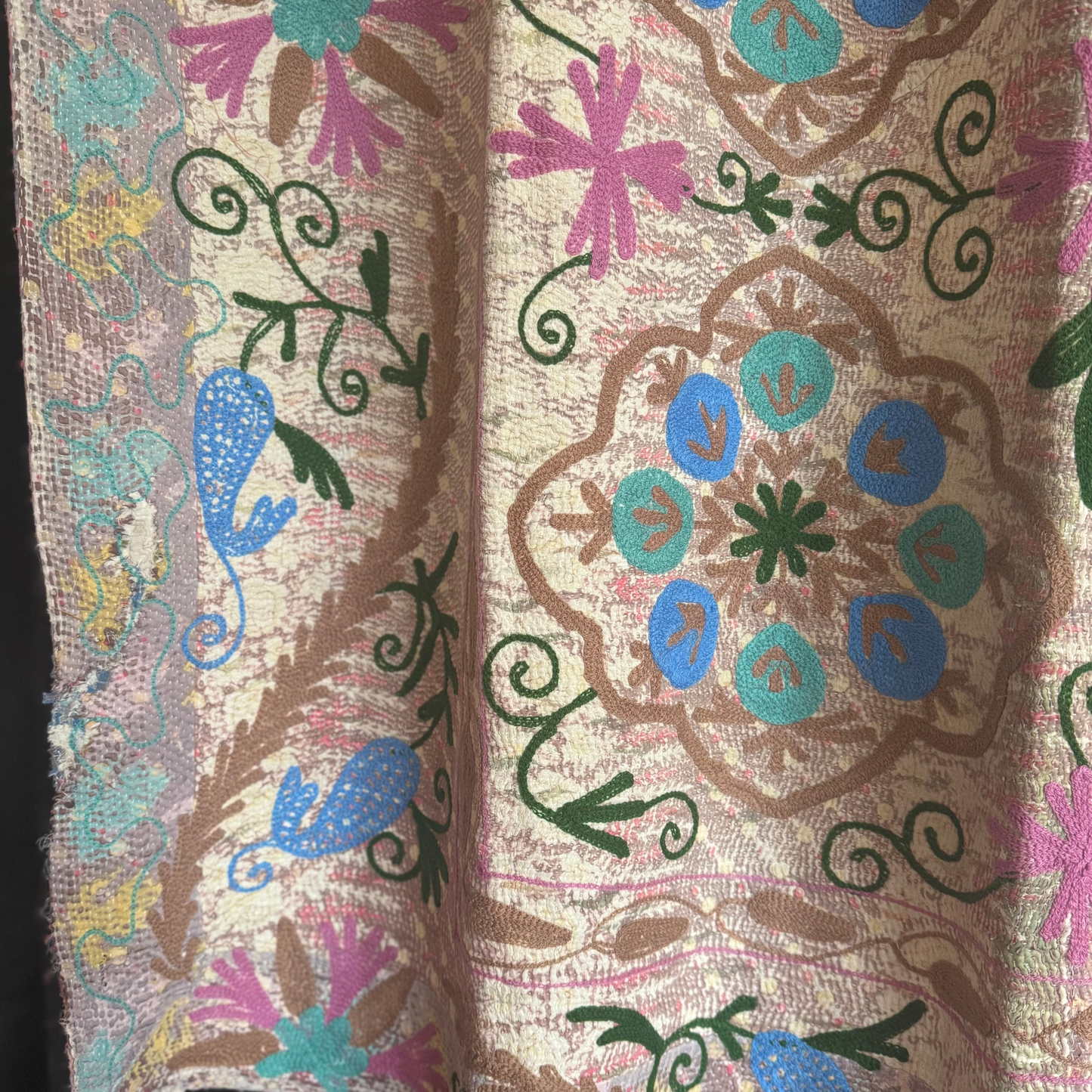 Close-up of hand-stitched turquoise and sky blue floral medallion framed by olive-green vines on a pastel-washed ground