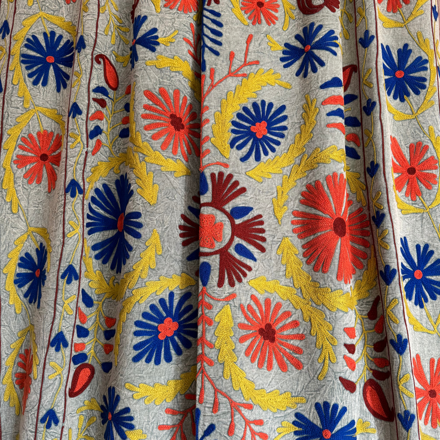 Close-up of dense chain-stitch embroidery in coral-red daisy and mustard-yellow vine on grey-green cotton
