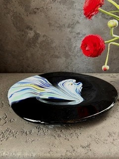 Close-up of a handcrafted Murano glaze plate with flowing swirls of blue, white, and gold against a deep black glass background.

